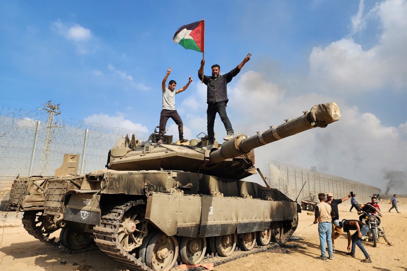 Members of Hamas' armed wing hold a Palestinian flag atop an Israeli tank. A border fence stretches behind them in the distance.