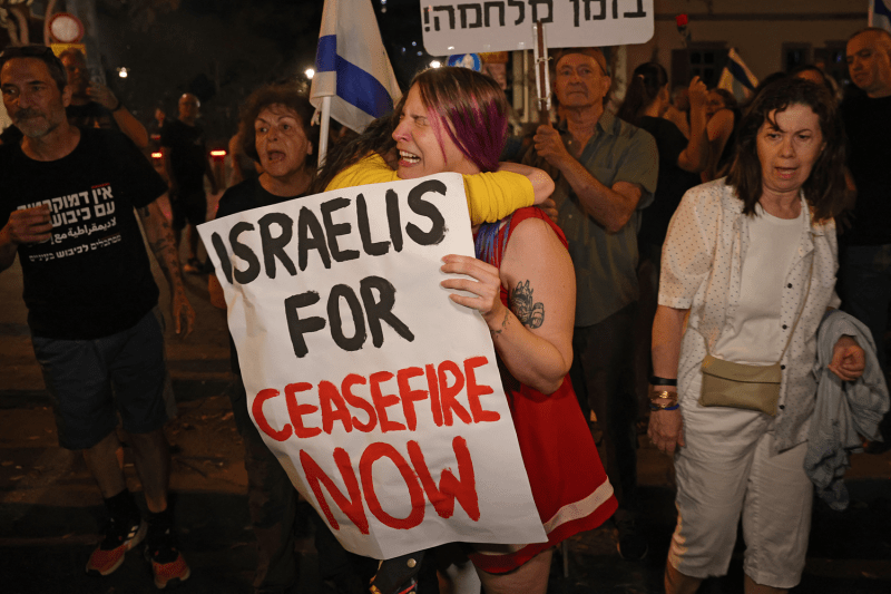 A group of protesters stand on a street at nighttime, many of them holding signs and chanting. In the foreground, a woman with brown hair dyed with purple streaks sobs as another demonstrator hugs her. The crying woman holds a hand-painted sign that says "Israelis for ceasefire now."