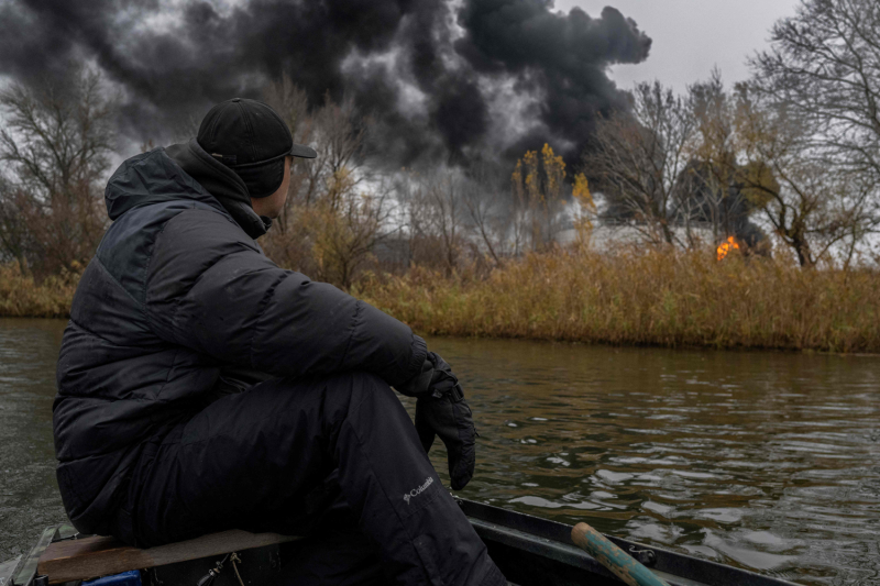 A fisherman wearing a heavy winter coat and hat can be seen from the side as he turns to watch a plume of smoke rising from a building beyond the far bank of the Dnipro. The man sits on a bench at the stern of his small railboat, and the sky above is overcast and pale gray.