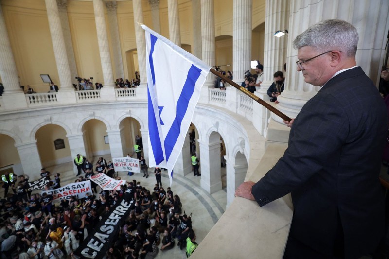 Republican Congressman Brandon Williams of New York holds an Israeli flag as he looks down on a crowd of protesters below as they stage a demonstration in support of a cease fire in Gaza. The crowd holds signs that say Ceasefire, Jews Say Ceasefire Now, and Philly Jews Say Never Again is Now. Reporters and photographers are seen on the columned balcony framing the scene in the Cannon House Office Building.