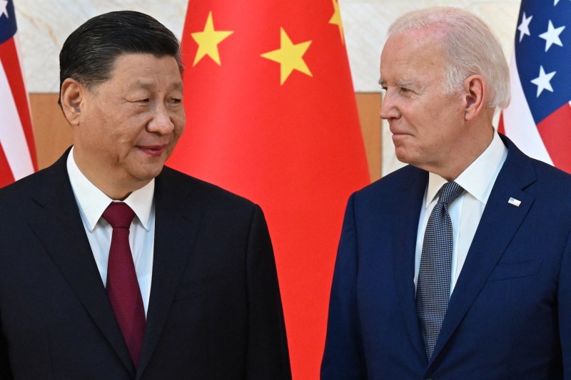 U.S. President Joe Biden and Chinese President Xi Jinping stand on the sidelines of the G-20 summit in Bali. U.S. and Chinese flags hang behind them.