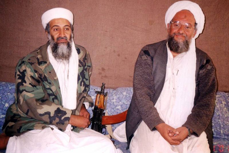 Bin Laden and Zawahiri sit side by side on the floor. A gun rests on the floor between them.