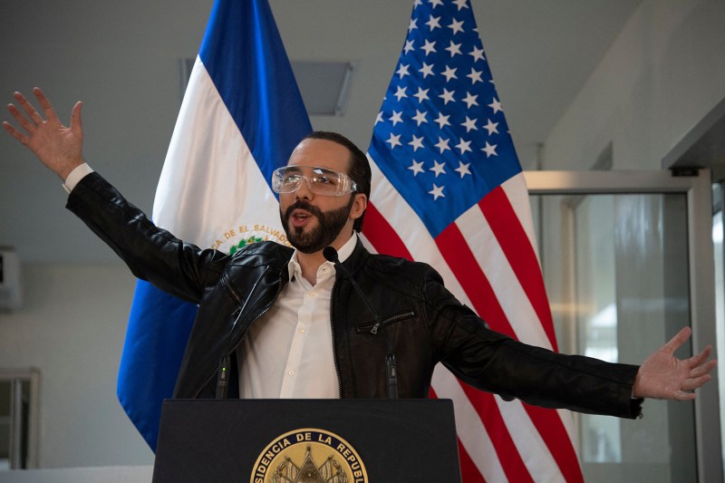 El Salvador's president Nayib Bukele, wearing clear plastic goggles and a black zip-up jacket, spreads his arms enthusiastically as he speaks during a joint news conference. He stands behind a podium and in front of U.S. and Salvadoran flags.