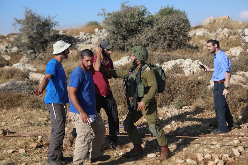 A group of man stand in a rocky field. An Israeli soldier wearing green military fatigues reaches out to push a hand against the chest of another man, who wears a red T-shirt and baseball cap as he gestures toward his own head. The man in red is flanked by two other men dressed casually in blue shirts, and all four seem to be speaking intensely.