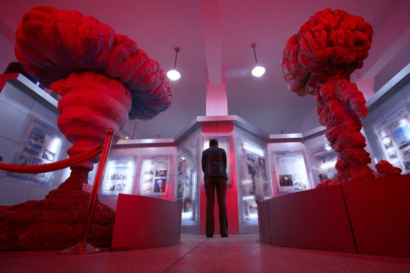 A man is seen from behind as he leans in to examine a glass case at an exhibit hall dedicated to a retired atomic bomb base in Tibet. The case includes photos and walls of text, and it is flanked on either side by two sculptures of mushroom clouds. The entire room is lit by dim, red-tinted light.