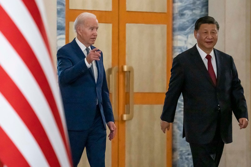 U.S. President Joe Biden, left, gestures as he walks past a U.S. flag. Ahead of him walks Chinese President Xi Jinping. Both wear suits as they walk past a large door.