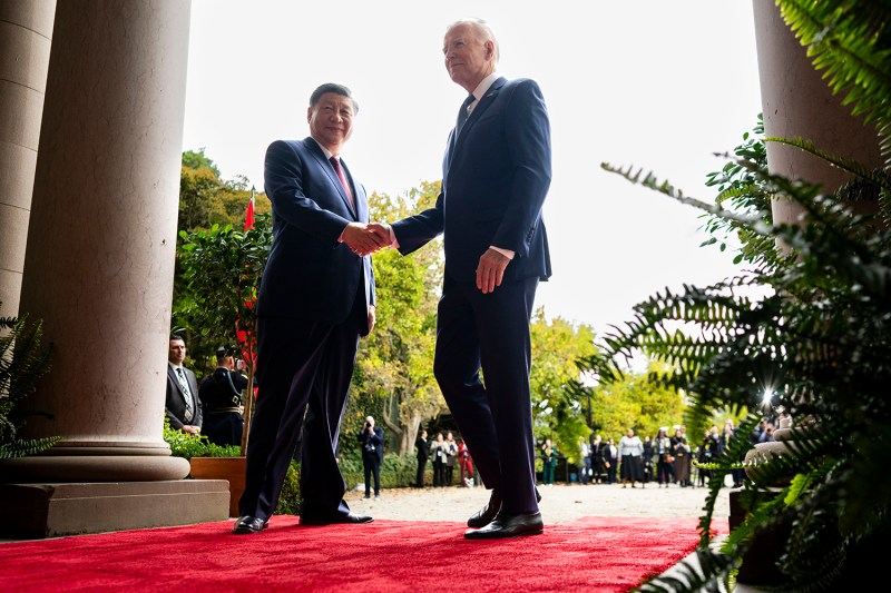 Biden and Xi shake hands while standing on a red carpet next to a white marble column.