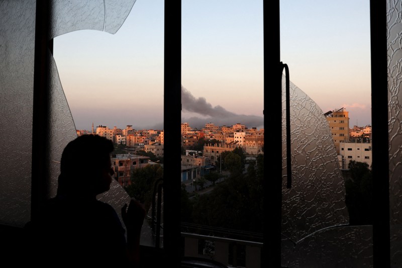 A Palestinian is seen from behind as they look outside through a large broken window. Some shards of fractured glass remain in the frame, but the rest of the opening reveals dark smoke billowing from a dense street of buildings in Gaza City. The sky is otherwise light but hazy.