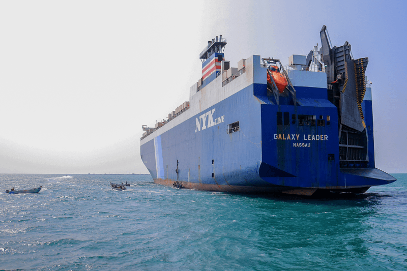 A massive container ship floats beneath a cloudy sky on the surface of the Red Sea near a port in Yemen. The hull of the ship is blue, and its name, the Galaxy Leader, is printed in white text on the stern.