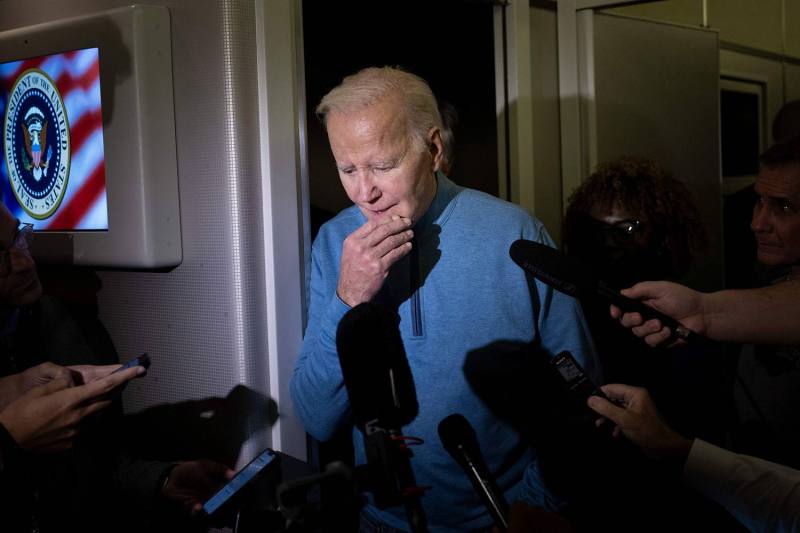 Biden, dressed in a casual half-zip sweatshirt, looks pensively downward as he stands facing numerous press microphones held out in his direction.