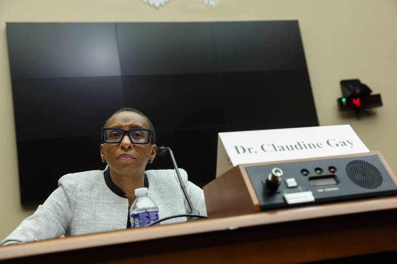 Gay is seen sitting behind a table with a desk microphone and a placard featuring her name.