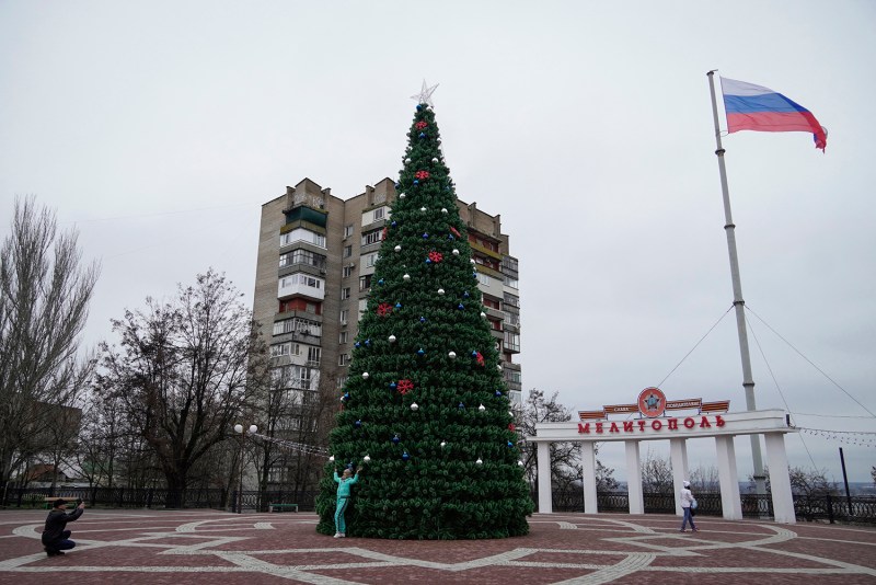 A woman poses for a photo in front of a tall decorated Christmas tree in front of a war-damanged building in Melitopol in Ukraine's Zaporizhzhia region with a Russian flag flying from a tall pole overhead.