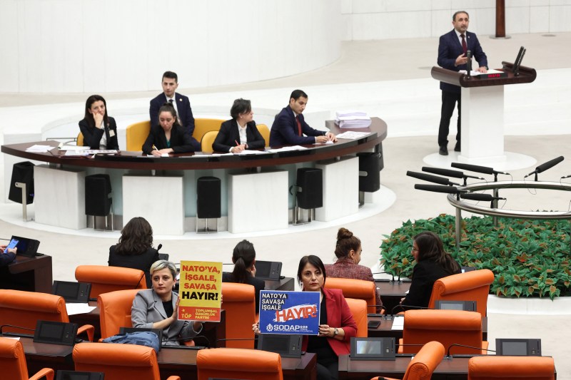 Two Turkish members of the opposition People's Equality and Democracy Party sit in the audience of a parliamentary meeting, facing back to the camera to hold up signs reading "No to NATO, Occupation, War" in the Grand National Assembly of Turkey.