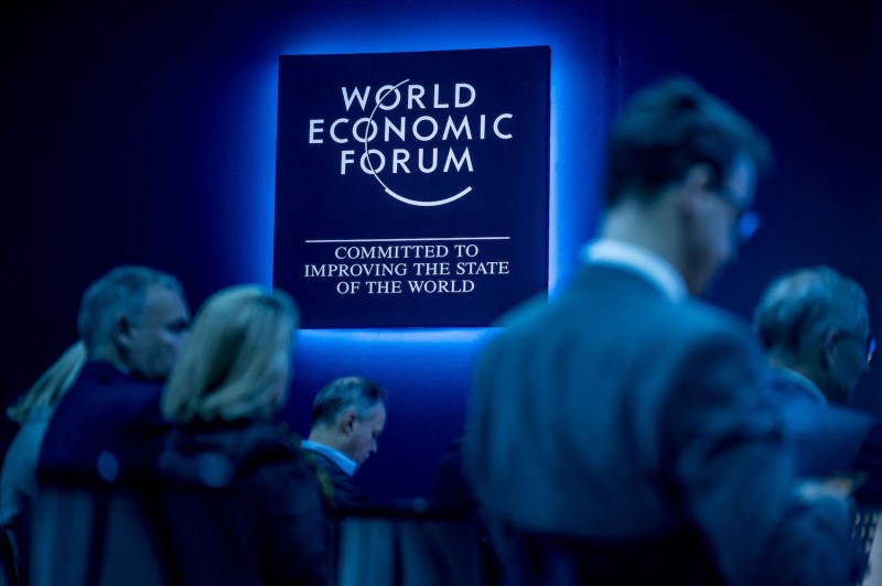 People sit in chairs before a glowing sign that reads: "WORLD ECONOMIC FORUM | COMMITTED TO IMPROVING THE STATE OF THE WORLD"