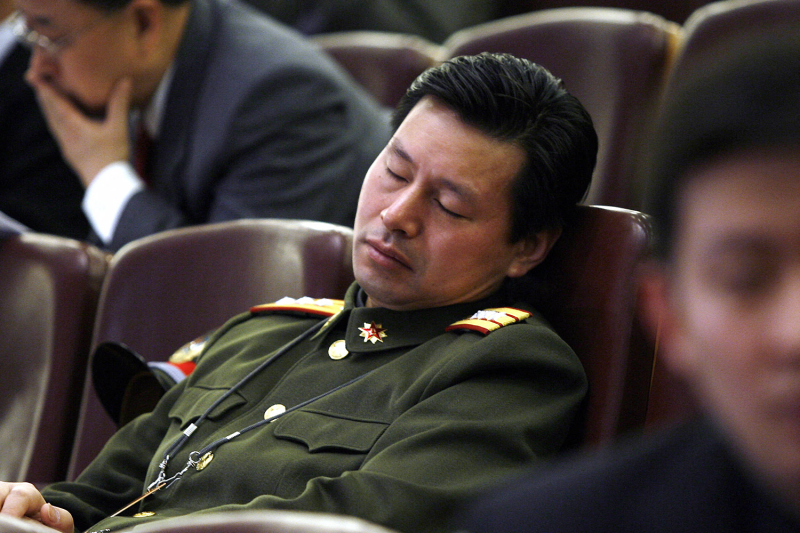 A People’s Liberation Army delegate wearing a dark green formal uniform with red and gold epaulettes sleeps while sitting in a chair at the Great Hall of the People. Other people in attendance at the congressional session sit in the rows of seats around him.