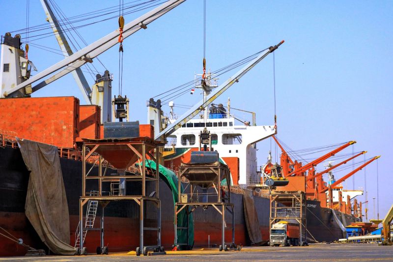 A wide shot shows a line of massive container ships moored at a port on the Red Sea in Yemen. Their cranes hang over the main platform of the port, and one is being used to lower a container into scaffolding above a truck.