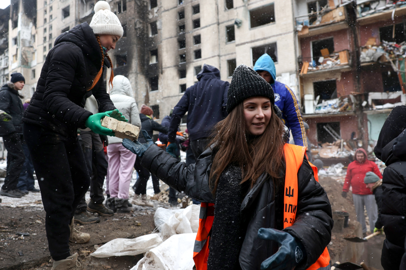 A woman wearing winter clothes and a reflective orange vest smiles as she hands a piece of rubble to another woman. Other volunteers stand around them as they help to clear rubble as snowflakes fall around them.