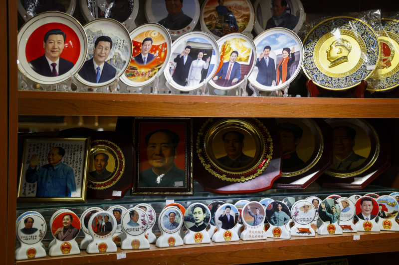 Collectible, decorative plates line a wooden display shelf. The ceramic plates are printed with various colorful images of Chinese leaders, including current Chinese President Xi Jinping and former communist party Chairman Mao Zedong.