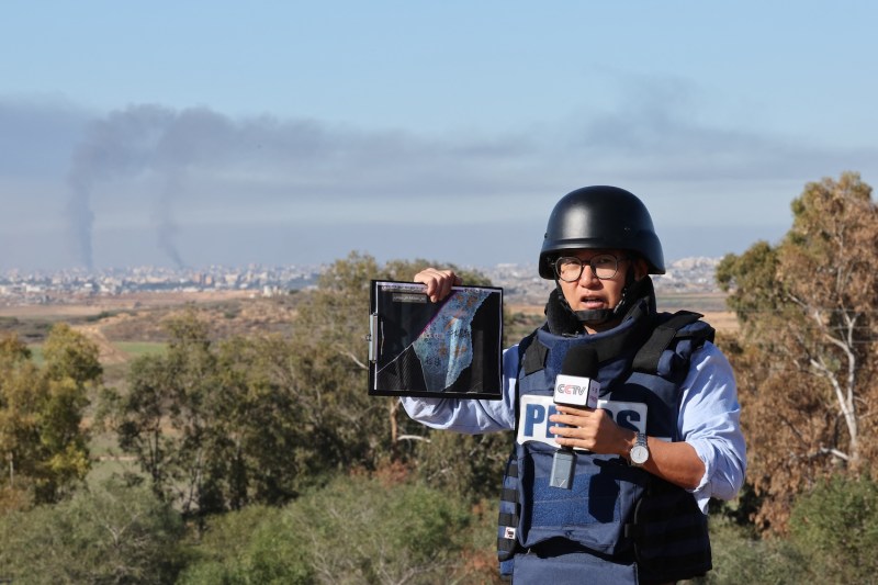 A Chinese journalist wearing a helmet and a bulletproof vest labeled "PRESS" speaks into a microphone while broadcasting for CCTV. In the background are fields and trees, and beyond that is the Gaza Strip, where columns of smoke from bombed buildings rise against a blue sky.
