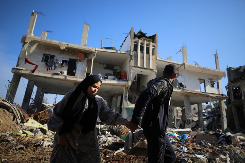 A woman wearing a headscarf holds hands with a man as they walk past a destroyed building in a refugee camp in the Gaza Strip. The man looks back over his shoulder at the devastated scene.