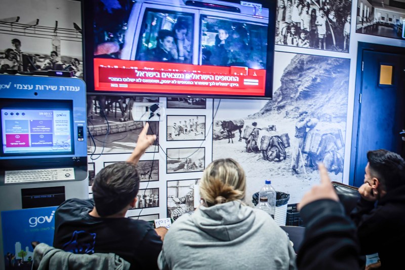 Three people wearing T-shirts and hoodies look up at a TV screen showing a news broadcast announcing the release of people from Hamas captivity. The walls of the kibbtz shelter where they sit are lined with historical photos.