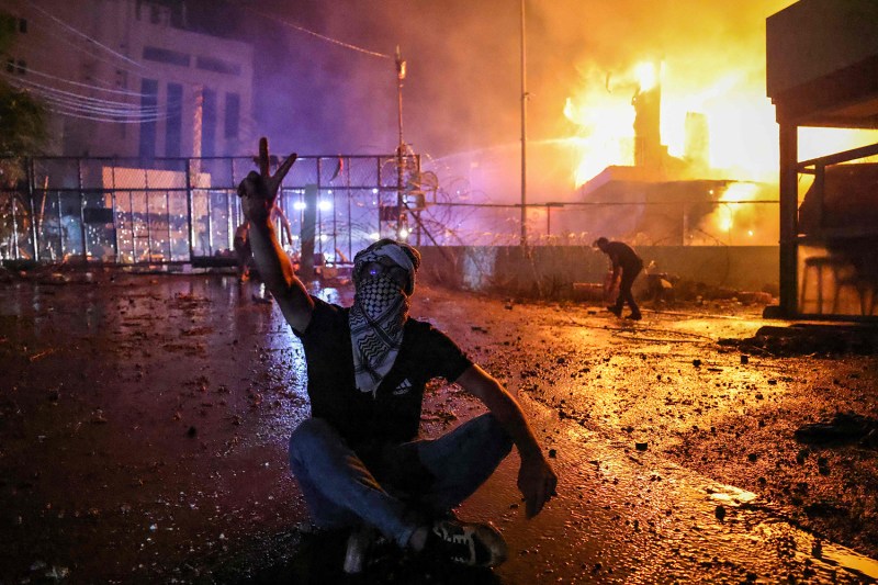 A protester sits cross-legged on the ground, wearing a keffiyeh, and flashes the victory sign. A fire rages behind the security gate of the U.S. embassy behind him. A person is seen behind him in the distance leaning over.