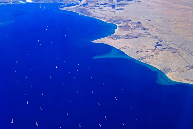 An aerial view shows stranded ships dotting bright blue water as they wait to cross the narrow Suez Canal seen in the distance at its southern entrance in the Red Sea.
