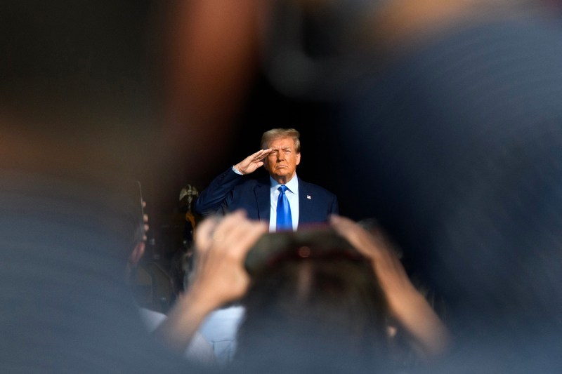 Former U.S. President Donald Trump, waring a suit and blue tie, salutes a crowd. The back of a person is seen holding up a phone to take a photo in the foreground.