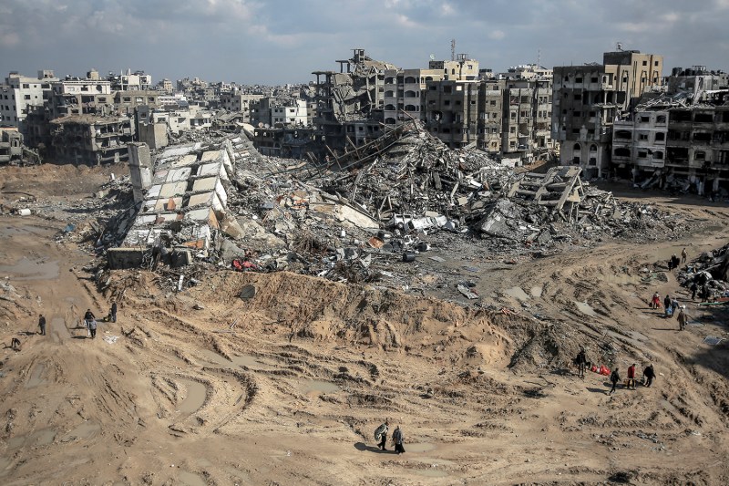 A dozen or so tiny figures are scattered across a bulldozed dirt landscape, picking their way around the rubble of a leveled building in the foreground. Devastated mid-rise towers stand in ruins in the distance with their windows blown out.