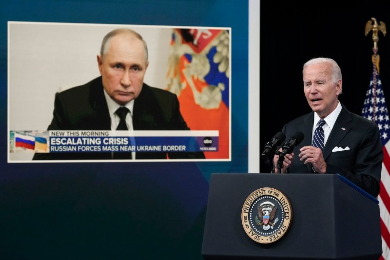 President Biden, dressed in a black suit, stands at a podium with an American flag behind him. To the side is a screen featuring the face and shoulders of Russian president Vladimir Putin.