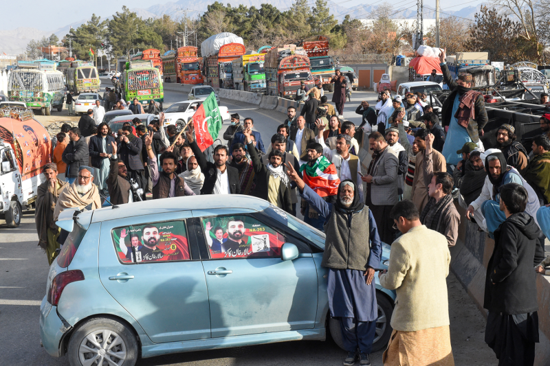 Supporters of imprisoned Pakistani politician Imran Khan surge onto a street as they block a highway to protest alleged election interference. Some demonstrators wave flags or hold up their hands. In the background, colorful trucks are backed up as far as the eye can see.
