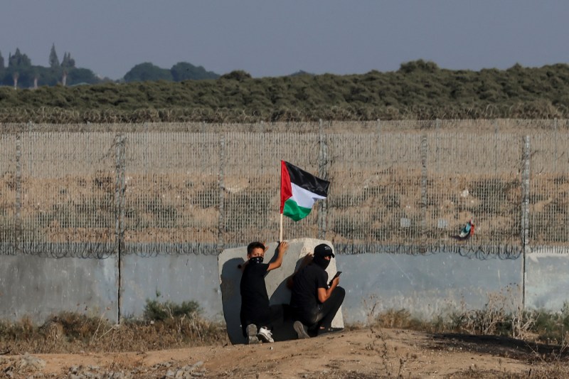 Two Palestinians, including a teenager, duck behind a slab of concrete as they wave the Palestinian flag next to the border fence of Gaza City. Scrubby grass and plants are visible in a field beyond the fence. Both people are dressed in black and wearing cloth face masks, glancing back over their shoulders at the camera.