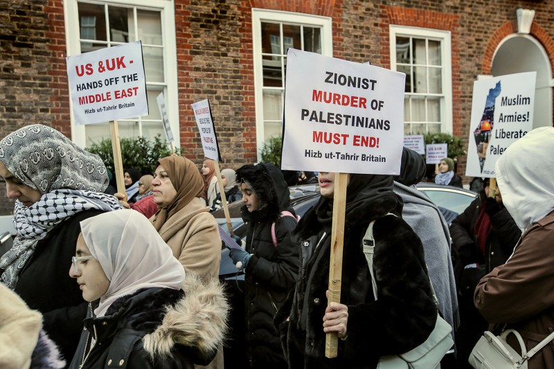 Women wearing headscarves and coats hold protest signs outside a brick building.