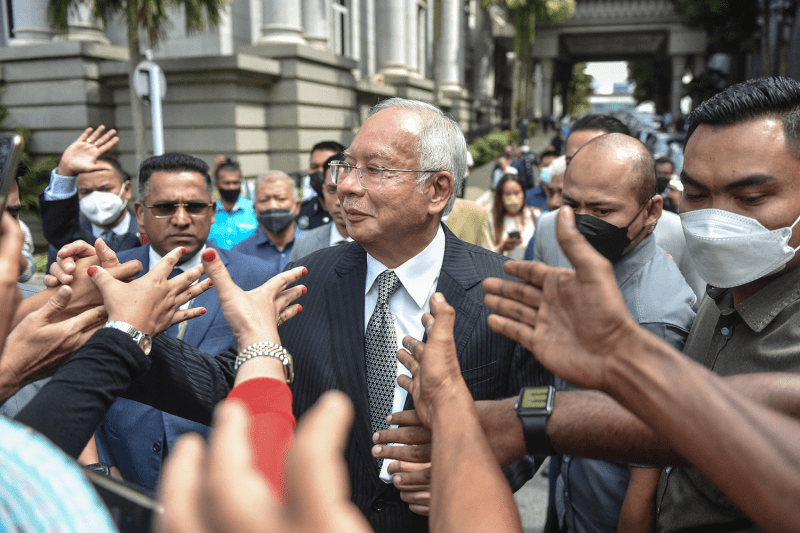 Former Malaysian Prime Minister Najib Razak stands in the middle of a crowd of supporters on the street, their hands reaching out to him as he smiles. Razak wears a suit and glasses as he stands outside a courthouse.