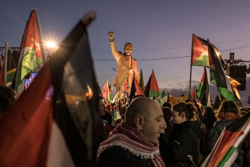 A crowd of people wave Palestinian flags under a dim sky at dusk as they gather around a statue of late South African President Nelson Mandela with his fist raised in the occupied West Bank city of Ramallah.
