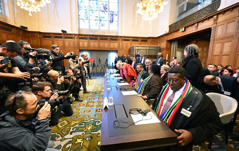 Several photographers and videographers swarm a bench of judges as they take their seats at the International Court of Justice in the Hague, the Netherlands. The room is lined with wooden paneling and chandeliers hang above. headphones and translation devices sit on the bench in front of the members of the court.