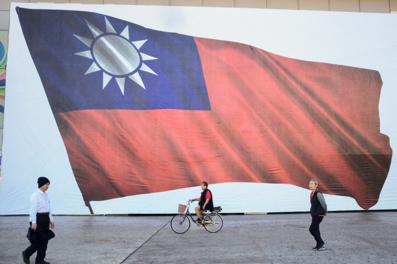 A giant depiction of the Taiwanese flag is seen on a street, with two people and a person on a bicycle going past it.