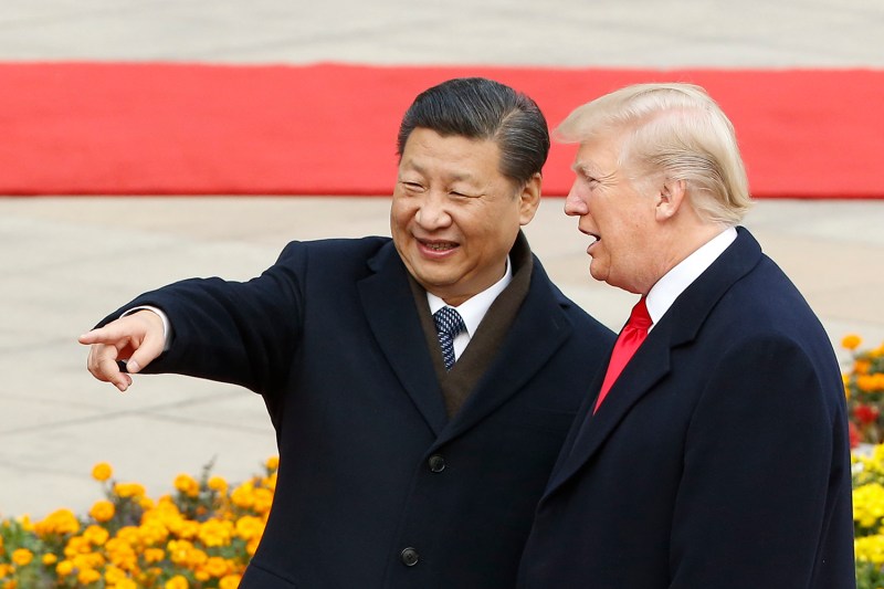 Chinese President Xi Jinping, wearing a dark overcoat, points and smiles as U.S. President Donald Trump talks to him. Behind them is a red carpet and flowers.