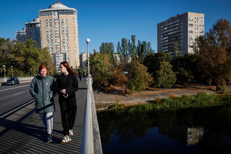 16-year-old Liza Batsura walks alongside her mother, Oksana, as they cross a bridge over a river in Kyiv. A few high-rise buildings stand against a deep blue sky behind them.