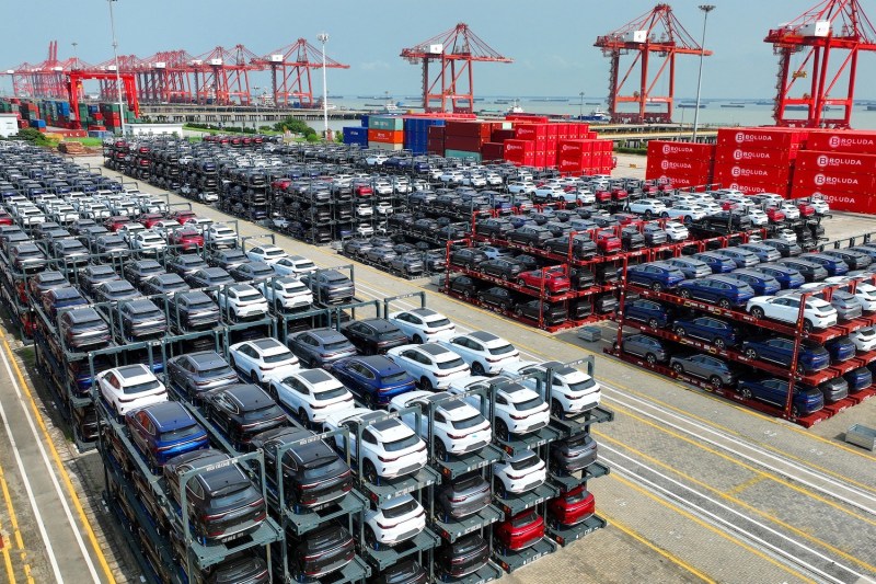 Hundreds of electric cars rest on large racks, each one with three layers of cars, beneath a smoggy sky at the container terminal of a port in China's eastern Jiangsu Province. Cranes and stacks of shipping containers loom in the background, and the ocean is visible beyond that.