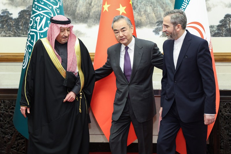 Wang Yi, a middle-aged man in a suit, puts his arms behind two other men standing on either side of him, one wearing a turban and robe, and the other in a suit, in front of flags of the countries of Saudi Arabia, China, and Iran.