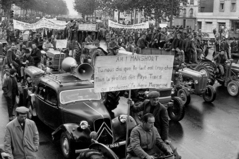 In black and white, a crowd of protestors and workers ride along an urban street in tractors and cars. The vast crowd reaches and disappears into the top left of the frame down the long street surrounded by trees on either side. In the near foreground a man riding in the front carriage of a car holds a tall sign that reads (in French): Ah ! Mansholt. You tell us that milk is too expensive but you benefit from third countries which produce the expensive margarine.