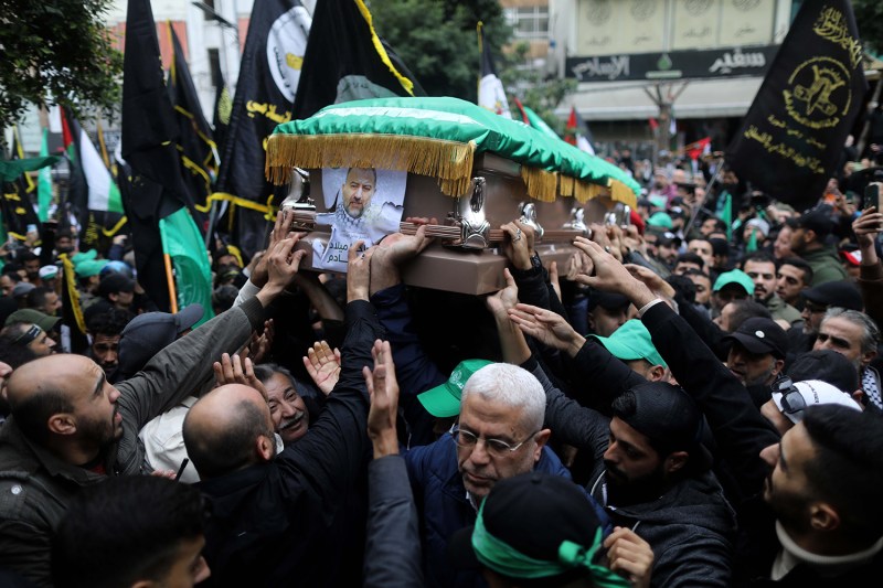 A crowd of people stretch their arms upward as several carry a flag-draped coffin with a picture of a man's face on it through the streets of Beirut.