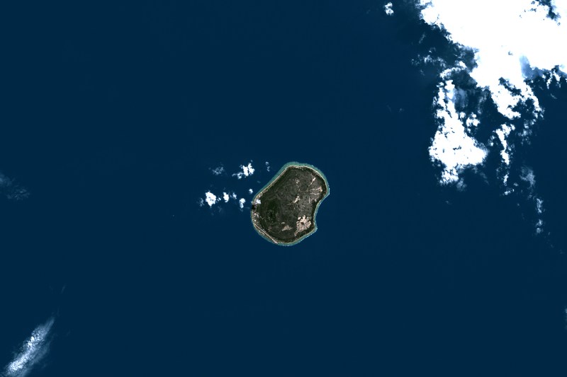 A satellite image shows the tiny island of Nauru in the South Pacific in June 1999. The dark blue ocean surrounds it, with a few clouds drifting into frame in the upper right corner.