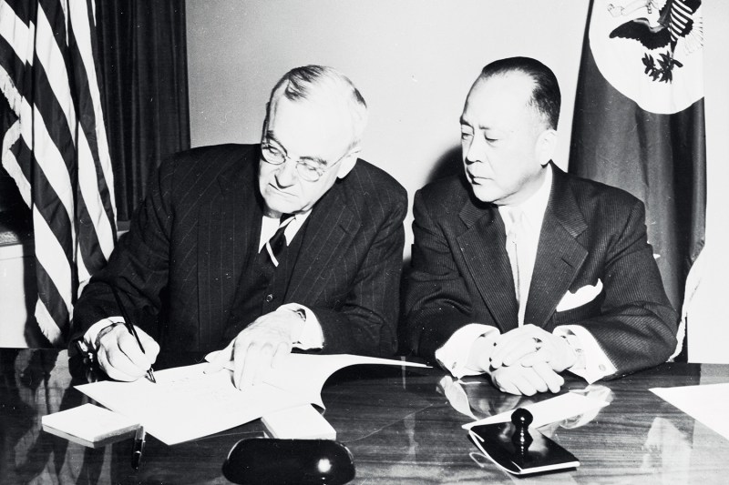 U.S. Secretary of State John Foster Dulles and Korean Ambassador You Chan Yang sit next to each other at a table as they sign a treaty. Both wear suits in a historical photo.