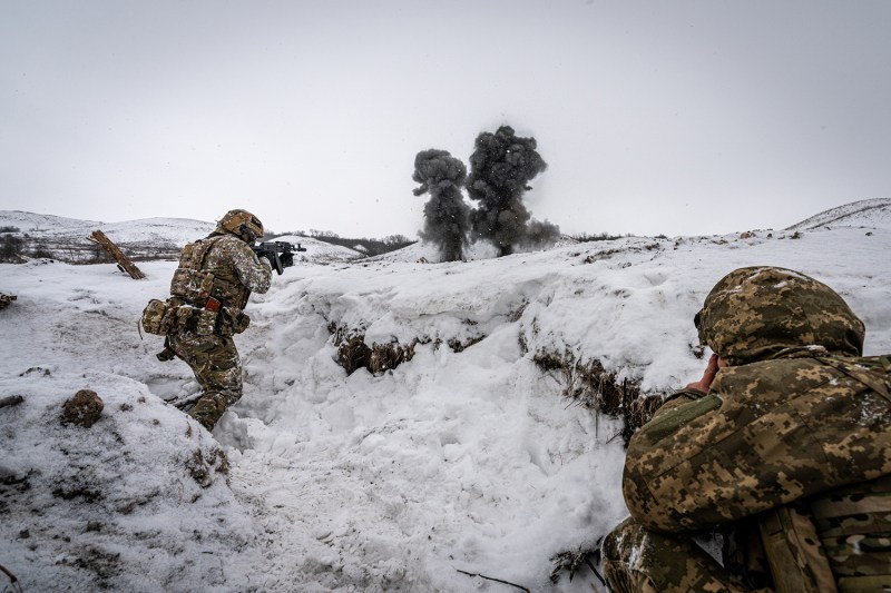 Two Ukrainian soldiers in uniform and helmets, with guns drawn move across a snowy landscape. Black smoke from an explosion is seen on the horizon.