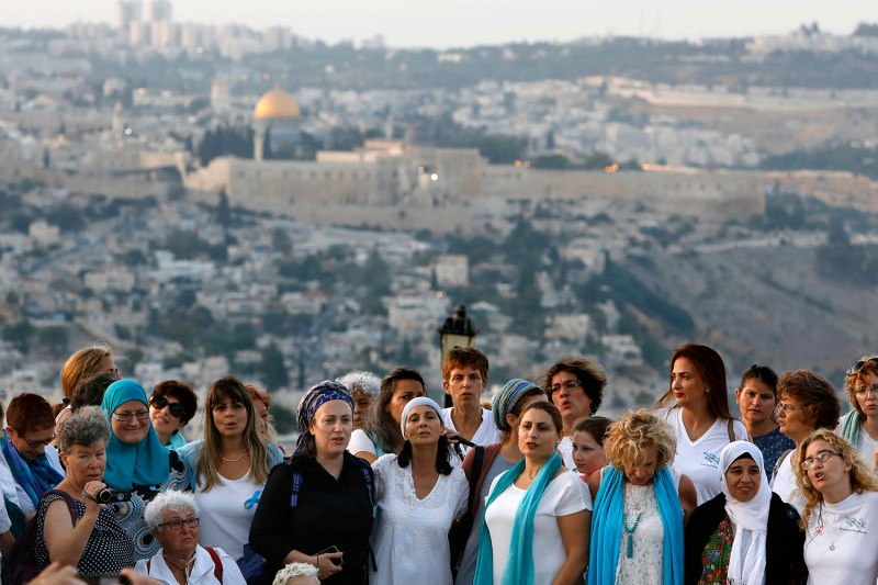 Dozens of women, wearing a mix of Jewish and Muslim scarves on their heads and shoulders, stand on a hill overlooking the Old City of Jerusalem.