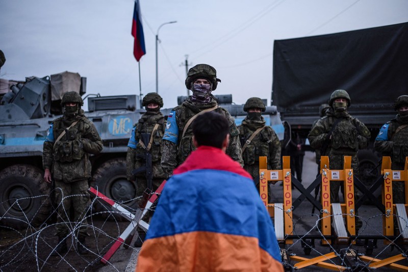 A protester wearing the Armenian flag over his shoulders like a cape is seen from behind as he faces a line of soldiers from a Russian peacekeeping mission. The shoulders wear camouflage uniform as well as masks that cover the lower halves of their faces, and they are separated from the protesters by a line of barbed wire.