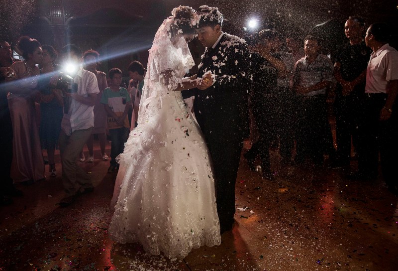 A bride in a white dress dances with a man in a suit during a wedding celebration at night, surrounded by guests. Confetti flutters through the air around the couple, some of it settling onto their shoulders.