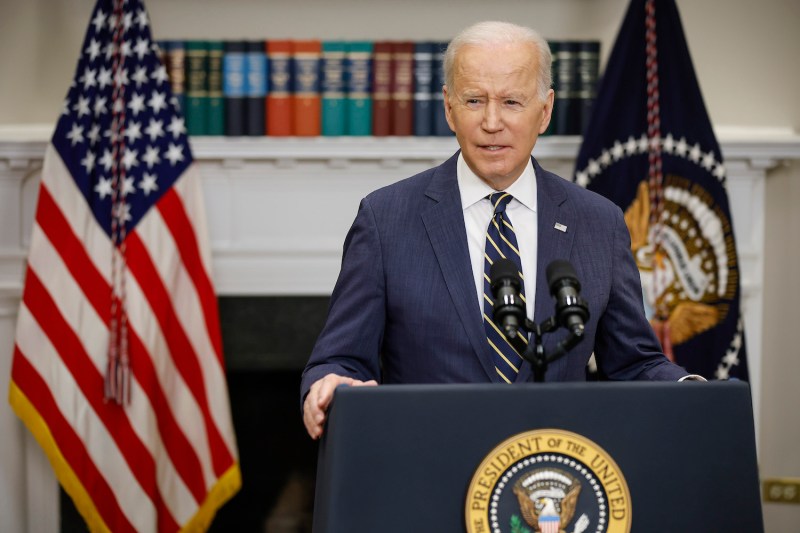 U.S. President Joe Biden speaks into a microphone while standing at a podium inside a room at the White House as he announces new economic actions against Russia. An American flag hangs behind him, in front of a mantle that holds a row of books.